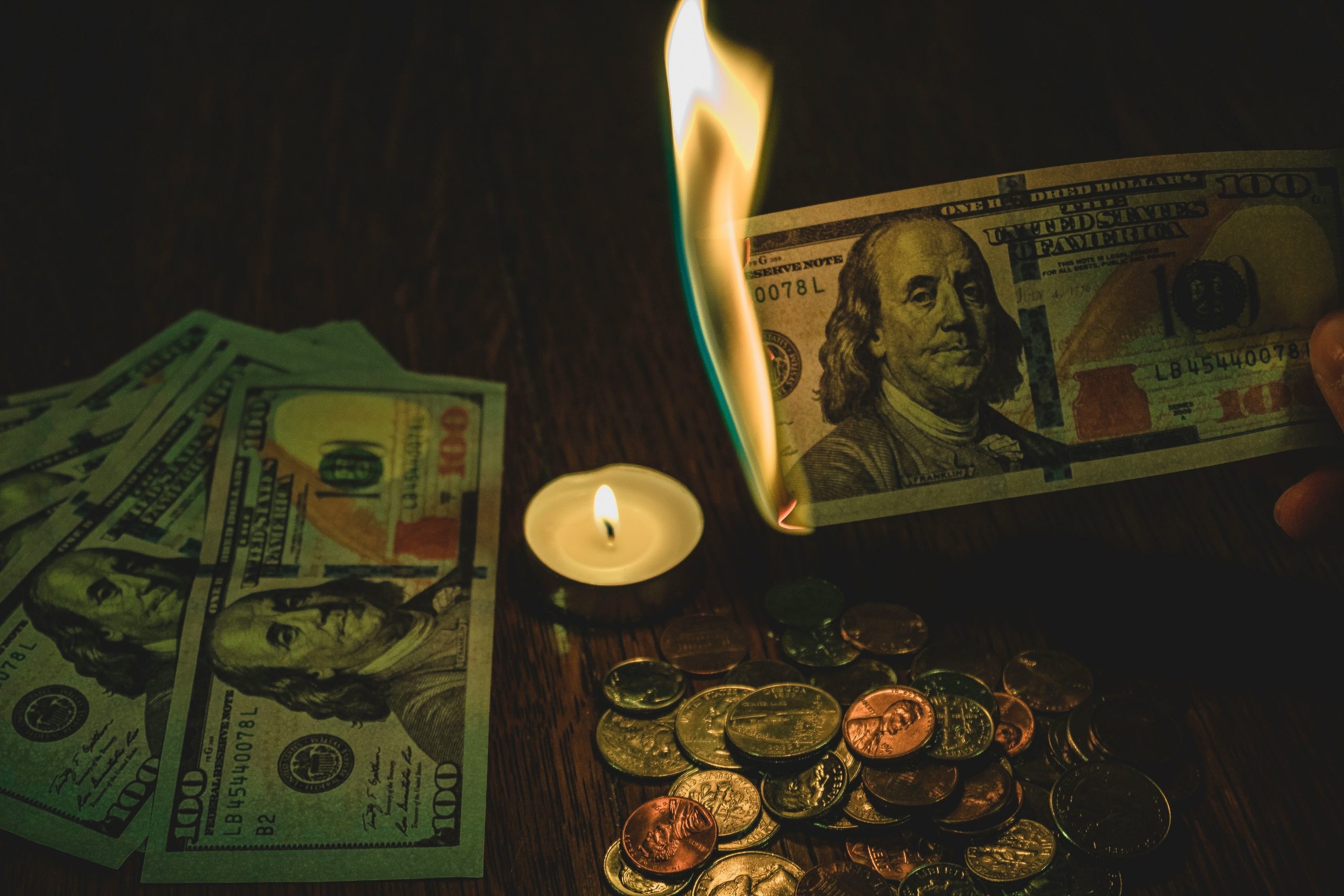 100 dollar bill in flames over candle surrounded by coins and 100 dollar bills.