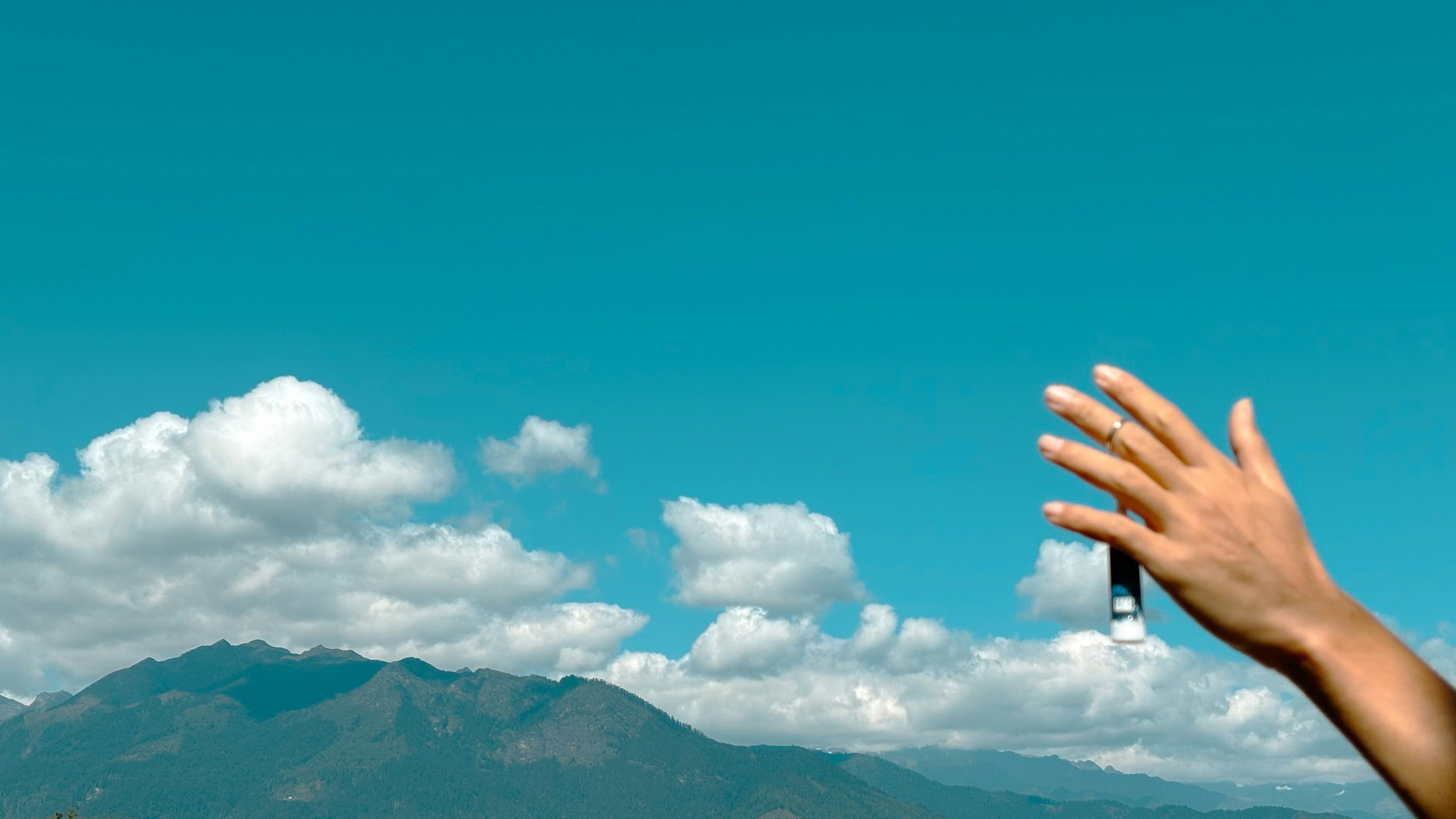 A hand reaches toward a vivid blue sky above distant green mountains and drifting clouds, creating a playful, airy composition that blends human presence with natural beauty.